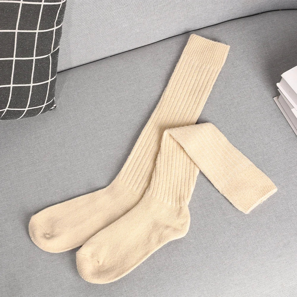 A pair of long, cream-colored ribbed socks crafted from a cashmere cotton blend is laid out on a light gray couch next to a black-and-white checked pillow and a stack of white books.