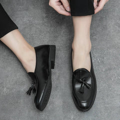 A person wearing black leather tassel loafers with black pants, sitting on a grey textured floor. One shoe, known for its cushioned insole, is on while the other foot is bare with the shoe held nearby.