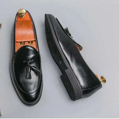 A pair of black leather loafers with tassels, displayed on a light surface. One shoe is upright with a shoe tree inside, while the other is laid on its side showing the sole and cushioned insole for added comfort.