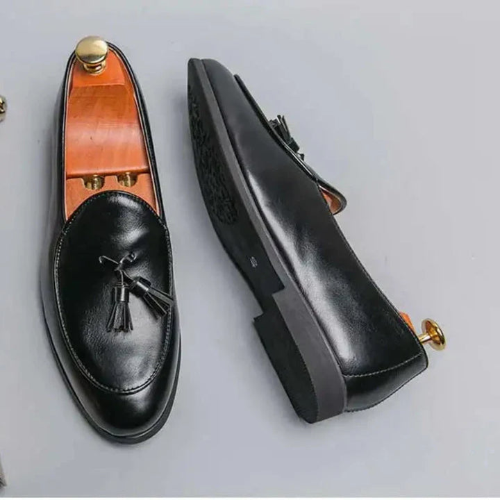 A pair of black leather loafers with tassels, displayed on a light surface. One shoe is upright with a shoe tree inside, while the other is laid on its side showing the sole and cushioned insole for added comfort.