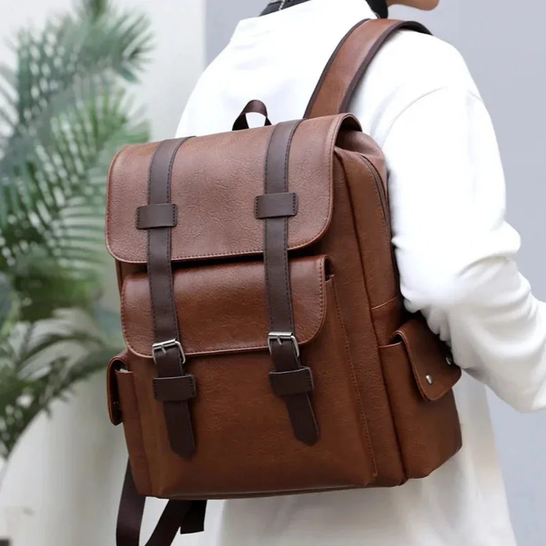 A person wearing a white shirt carries a large brown waterproof leather backpack with buckle straps and spacious compartments on their back. The background includes a green leafy plant.