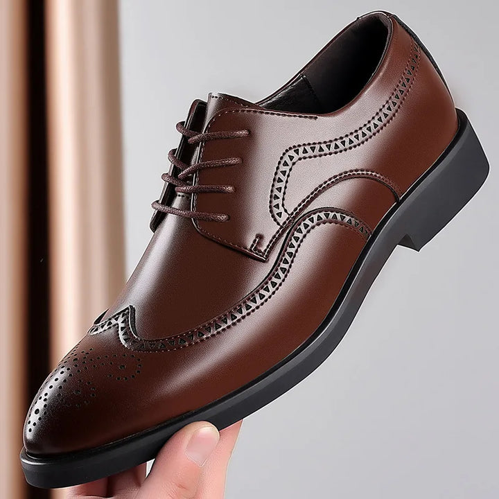 A hand holding a brown premium leather dress shoe with brogue detailing, black laces, and a black sole, against a plain background.