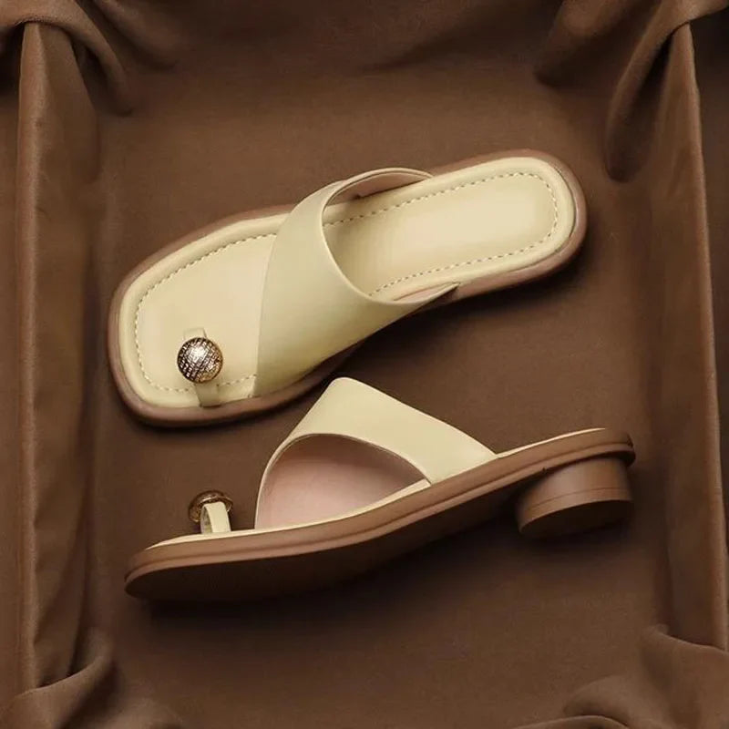 Two beige Square Toe Slides with low heels sit on a brown fabric surface. One sandal has a decorative round metallic accent near the toe area. These Women's Modern Slippers feature a minimalist, open-toe design.