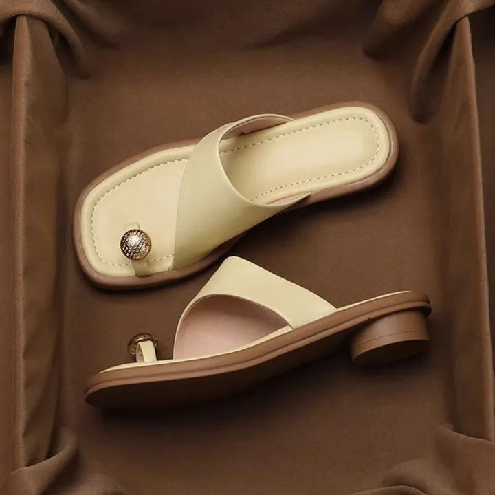 Two beige Square Toe Slides with low heels sit on a brown fabric surface. One sandal has a decorative round metallic accent near the toe area. These Women's Modern Slippers feature a minimalist, open-toe design.