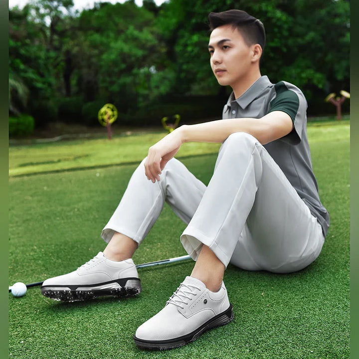 A man in a gray polo shirt and light pants sits on a grassy golf course, resting with one arm on his knee. Nearby, his high-traction outsole shoes and golf gear lie on the ground, framed by trees in the background.