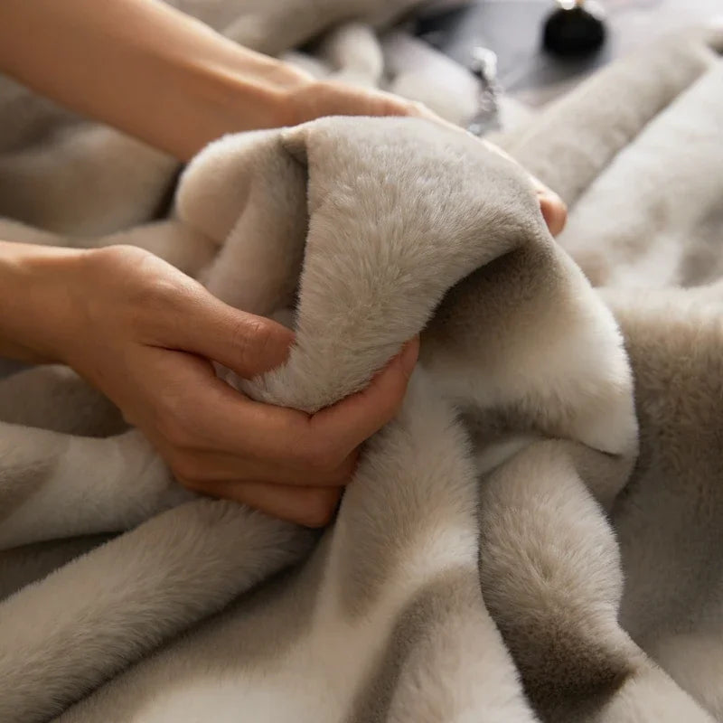 Two hands gently hold and squeeze a soft, plush, beige and white faux rabbit fur blanket, highlighting its smooth and fluffy texture.