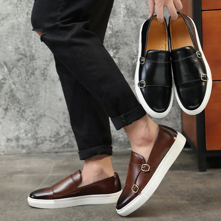 A person in black pants wears brown Slip-On Loafers with white soles and gold buckles, holding a similar pair in black. The scene highlights British Fashion against a gray wall and a wooden cabinet.