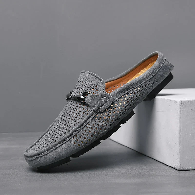 A single grey suede loafer with perforated detailing, innovative cushioning, a black sole, and a decorative metal accent on top, displayed against a grey background and resting partially on a white block.