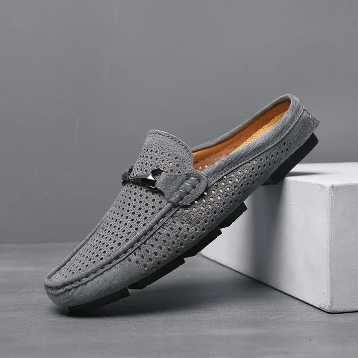 A single grey suede loafer with perforated detailing, innovative cushioning, a black sole, and a decorative metal accent on top, displayed against a grey background and resting partially on a white block.
