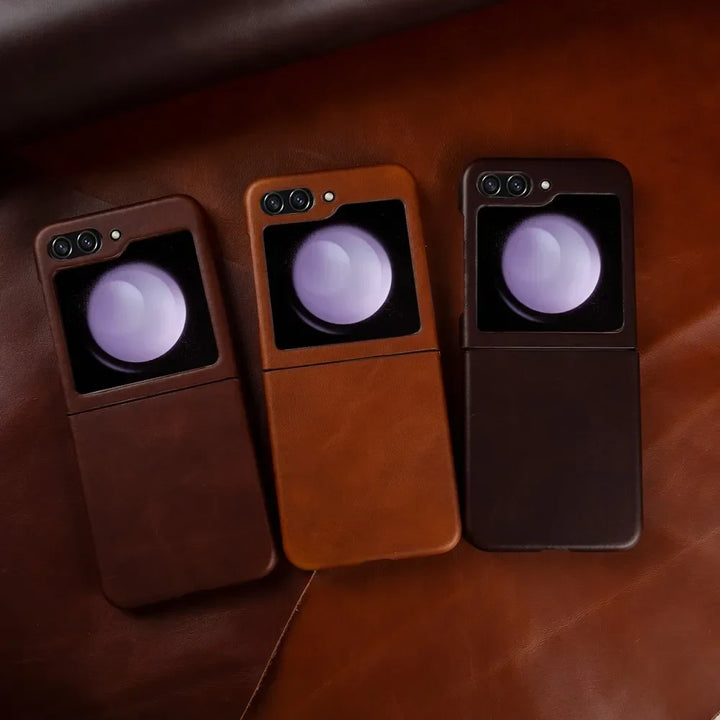Three Samsung Galaxy Z Flip smartphones in genuine leather cases are placed side by side on a brown leather surface, each phone displaying a purple circular reflection on its external screen.