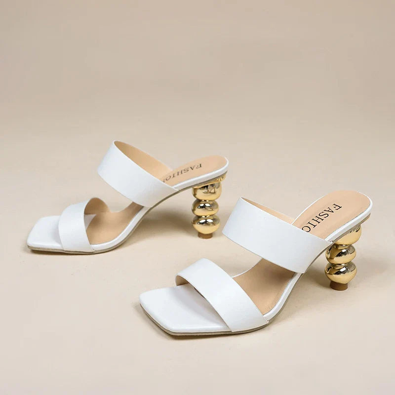 A pair of white high-heeled sandals with square toes, double straps, and unique gold sculpted heels, crafted from premium leather and placed on a plain beige background.