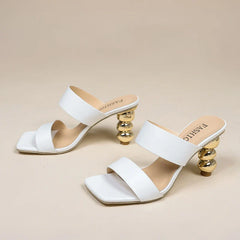 A pair of white high-heeled sandals with square toes, double straps, and unique gold sculpted heels, crafted from premium leather and placed on a plain beige background.