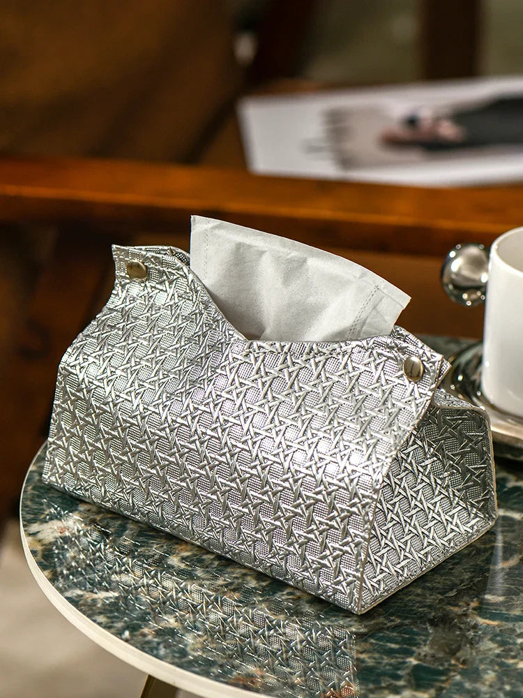 A silver Glymmer Tissue Box with a geometric pattern sits on a marble table, holding white tissues. A white cup and saucer and a magazine are visible in the background.