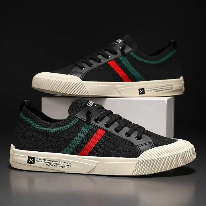 A pair of black low-top sneakers with a breathable mesh upper, white soles, green and red stripes on the sides, black laces, and white text details above the sole, displayed against a dark background.
