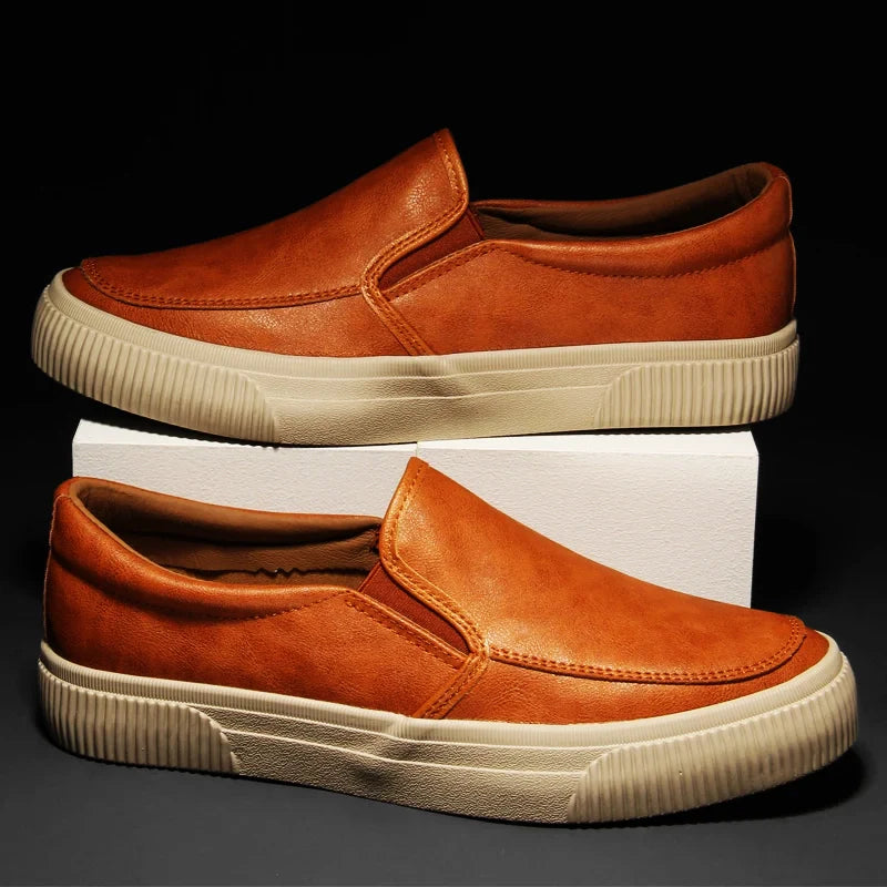A pair of brown men’s slip-on leather sneakers with beige hybrid rubber soles are displayed, one shoe resting on a white rectangular block and the other placed flat in front, against a black background.