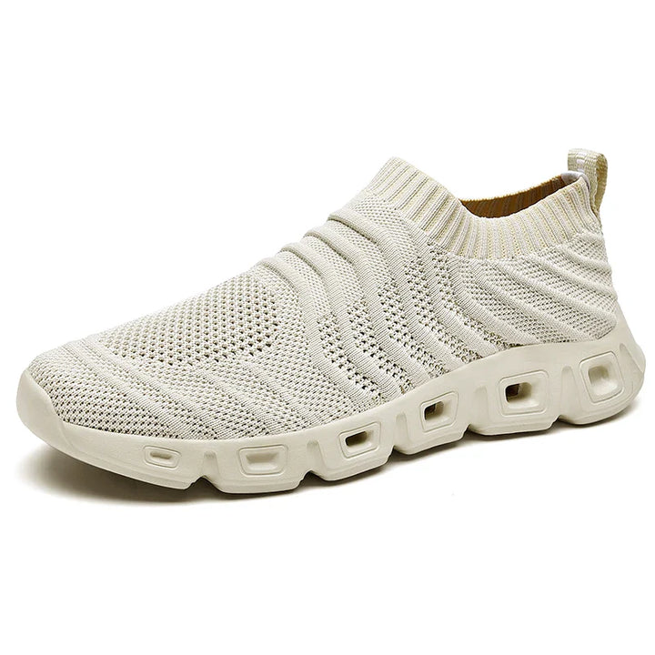 A light beige athletic slip-on shoe with a knitted, breathable upper and a cushioned sole featuring square-shaped cutouts along the sides, designed for a sock-like fit.