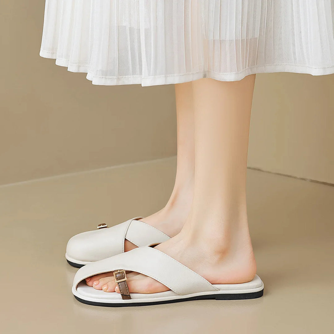 A person wearing a white pleated skirt and chic leather slides with crisscross straps and a small buckle, standing on a beige floor—perfect for an elegant summer wardrobe.