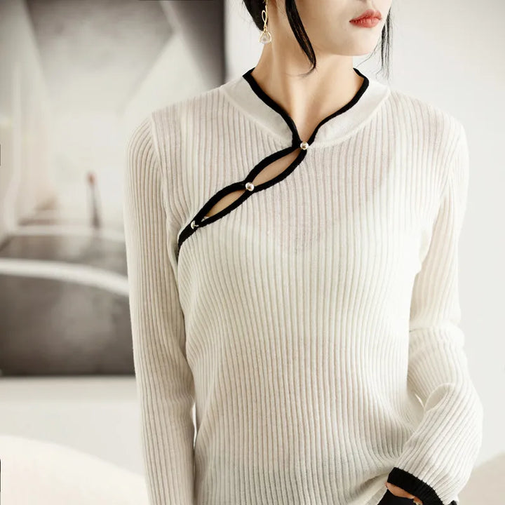 A woman wears a ribbed knit white long-sleeve top with a unique diagonal keyhole design accented by black trim and decorative buttons. She has short dark hair and is wearing a single dangling earring.