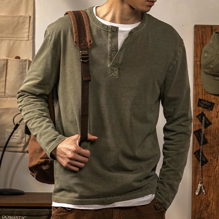 A person wearing a casual olive green pure cotton long-sleeve Henley shirt carries a brown shoulder bag. Only the torso and part of the head are visible. The background includes a cap, wall organizer, and wooden panel.