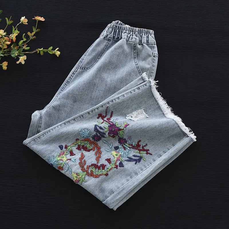 Light loose-fitting denim jeans with an elastic waistband, folded neatly on a dark surface. One leg features retro embroidery and beadwork in floral and leafy patterns. Small leaves or flowers sit at the top left corner.