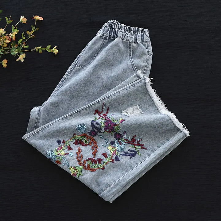 Light loose-fitting denim jeans with an elastic waistband, folded neatly on a dark surface. One leg features retro embroidery and beadwork in floral and leafy patterns. Small leaves or flowers sit at the top left corner.