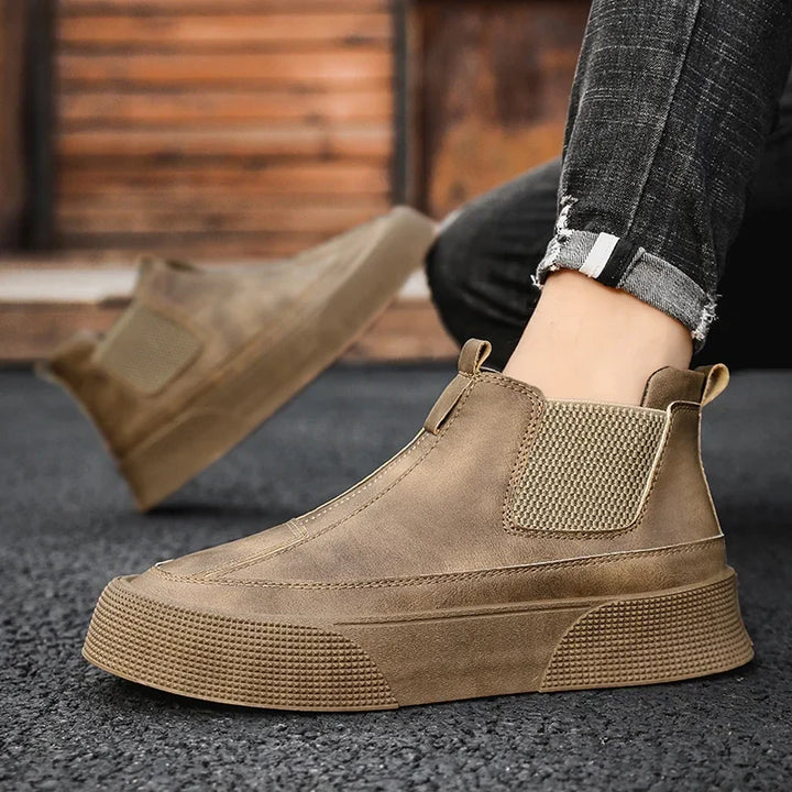 A person wearing brown vintage high-tops with textured soles, elastic side panels, and a cushioned footbed, paired with rolled-up dark jeans, stands on a blacktop surface against a wooden background.