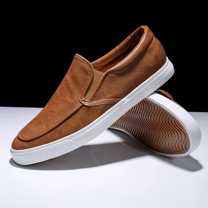 A pair of brown mens slip-on casual shoes with white rubber soles and a comfortable leather insole, one standing upright and the other leaning against it, displayed on a flat white surface with a dark background.