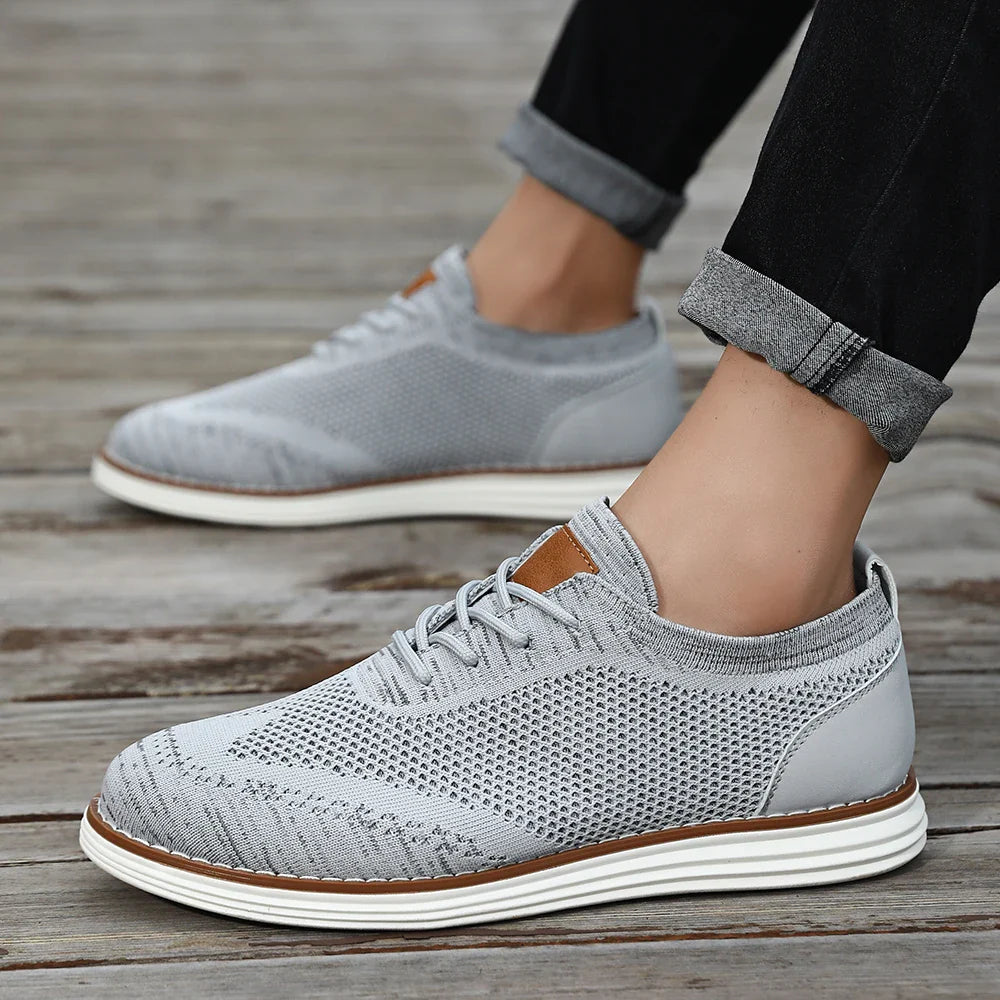 Gray lightweight sneakers with white soles are worn by someone standing on a wooden surface. The person is sporting black pants with rolled cuffs, showing off their ankles for a relaxed, casual style.