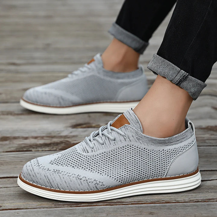 Gray lightweight sneakers with white soles are worn by someone standing on a wooden surface. The person is sporting black pants with rolled cuffs, showing off their ankles for a relaxed, casual style.
