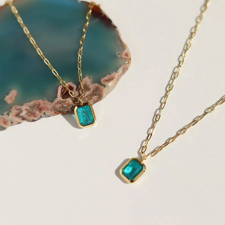 Two 18K gold plated chain necklaces with rectangular blue-green gemstone pendants are displayed on a white surface, with part of a teal and brown agate slice in the background. These pieces feature hypoallergenic materials for sensitive skin.