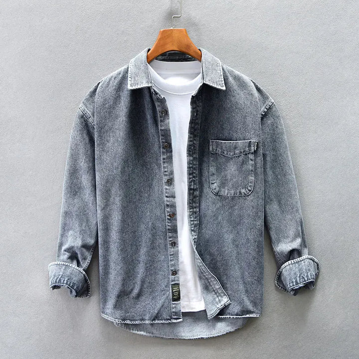 A washed denim button-up overshirt with rolled-up sleeves is layered over a plain white T-shirt and displayed on a wooden hanger against a light gray background.