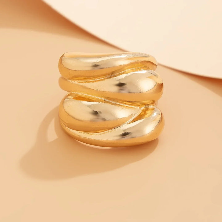 A chunky, gold-tone ring with a smooth, wavy, layered geometric design is displayed on a beige surface with a soft shadow, creating a stylish and elegant appearance.