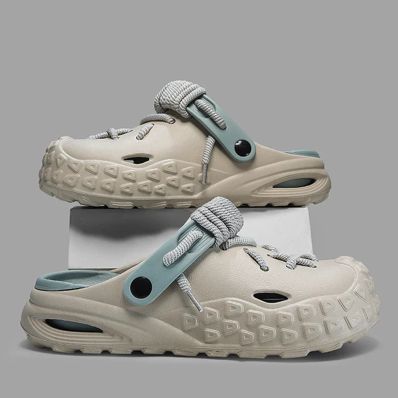 A pair of beige slip-on clogs with textured soles for enhanced grip, blue-gray adjustable straps, and gray rope laces with lace detail, displayed stacked on a white platform against a plain gray background.