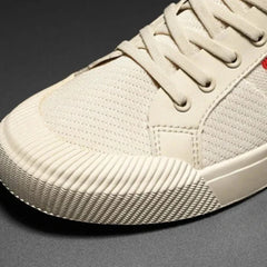 Close-up of the front and side of a beige sneaker with a textured rubber toe cap, canvas material, and matching beige laces, featuring a durable rubber outsole, photographed against a dark background.
