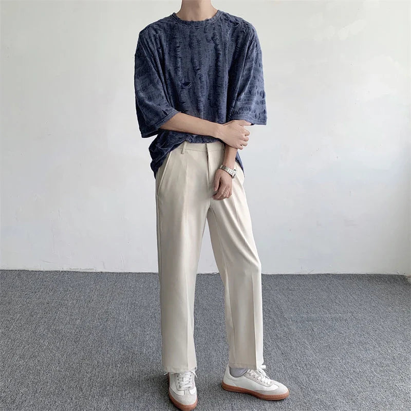 A person stands indoors against a plain wall, wearing an oversized dark blue streetwear tee, light beige trousers, and white sneakers, with arms casually crossed. Their head is not visible in the image.