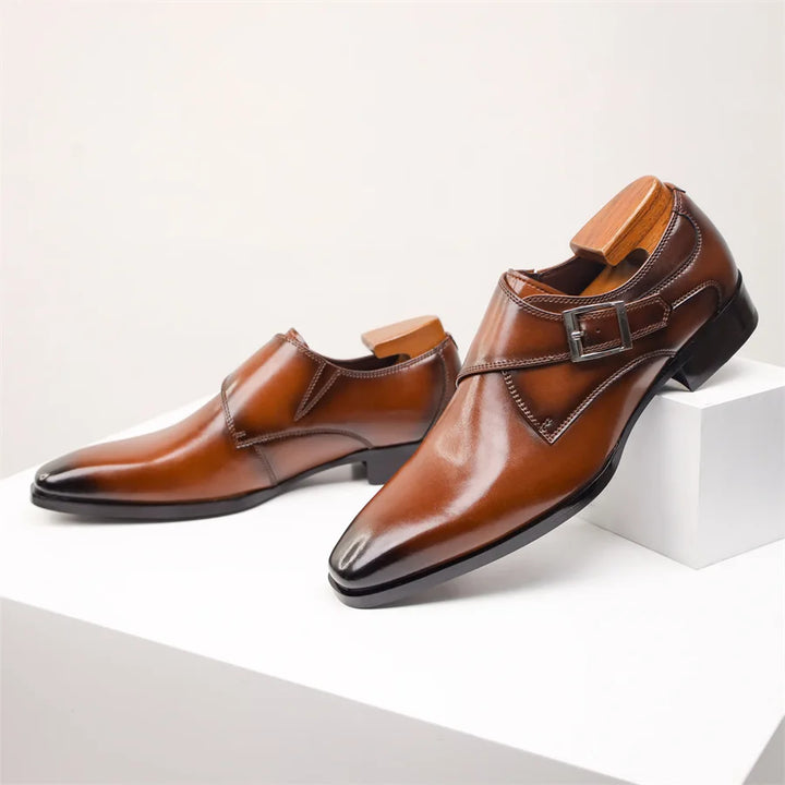A pair of polished brown Italian leather monk strap shoes with a side buckle detail, displayed on a white surface with wooden shoe trees inside each shoe.