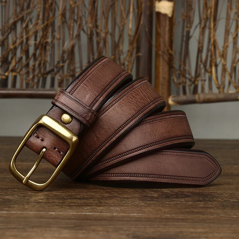 A genuine leather cowhide belt with a brass buckle is coiled on a wooden surface, accented by decorative branches and a rustic background.