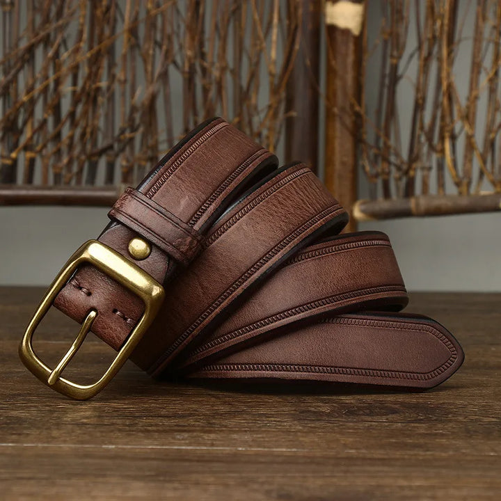 A genuine leather cowhide belt with a brass buckle is coiled on a wooden surface, accented by decorative branches and a rustic background.