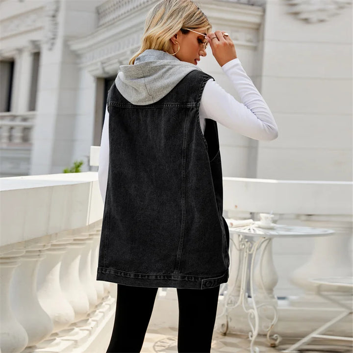 A woman with blonde hair, wearing sunglasses, a white long-sleeve shirt, a gray hood, and a lightweight breathable sleeveless denim vest, stands on an elegant white balcony with classic architectural details.