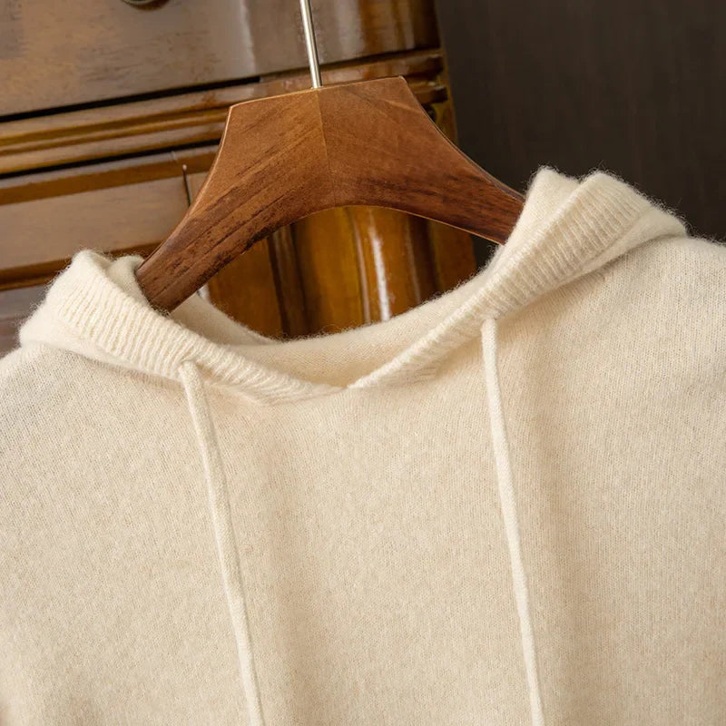 A close-up of a cream-colored, soft knit cashmere blend hoodie on a wooden hanger, with drawstrings and ribbed edges, hanging in front of a wooden dresser.