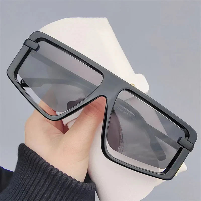 A hand holds a pair of oversized black rectangular sunglasses with dark tinted UV400 lenses, offering superior sun protection, against a plain light gray background.