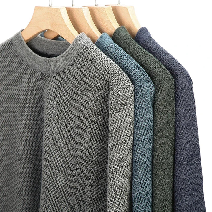 Four textured knit sweaters in light gray, teal, dark green, and navy blue—each a high-quality cotton sweater—hang neatly on wooden hangers against a white background.