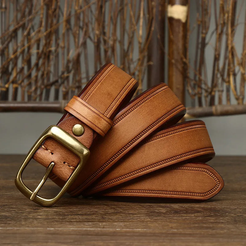 A tan cowhide belt with a brass buckle is coiled on a wooden surface, with a rustic, twig-style background visible behind it.