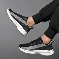 A person wearing black and gray workout shoes with breathable mesh, featuring white soles and black pants, lifting both feet against a plain gray background.