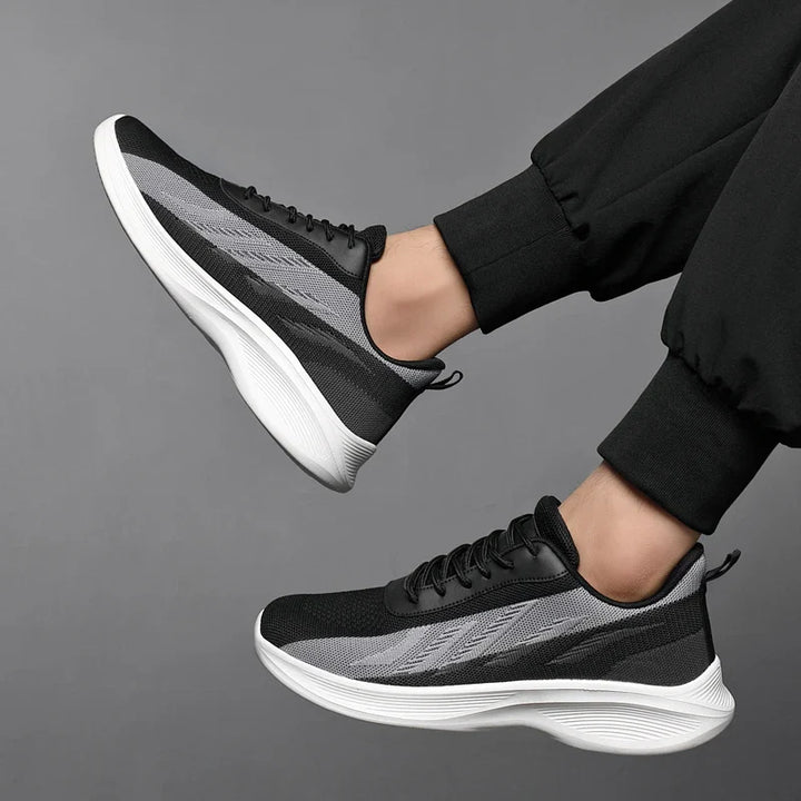 A person wearing black and gray workout shoes with breathable mesh, featuring white soles and black pants, lifting both feet against a plain gray background.