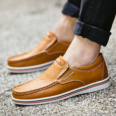 A person wearing light brown leather boat-style loafers with white and red-striped soles stands on a gravel surface. The person’s jeans are rolled up at the ankles.