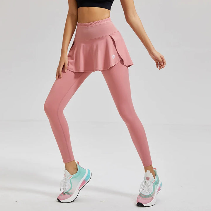 A person wearing a black crop top, pink skirted leggings with high-waisted support, and white sneakers with colorful accents stands in a minimal, light-colored studio.