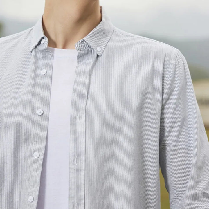A person wearing a classic pinstripe cotton button-up shirt over a white t-shirt, photographed from the shoulders to the chest, with an outdoor background blurred.