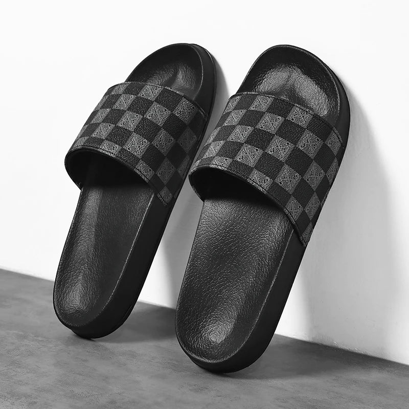 A pair of black checkered sandals with a textured footbed and a wide strap featuring a dark checkerboard pattern, leaning against a white wall on a gray floor—perfect streetwear slides for casual style.