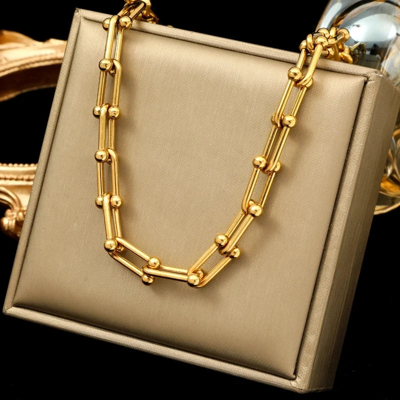 A gold statement chain necklace with elongated links and small round beads, crafted from 316L stainless steel, is displayed on a beige jewelry stand against a black background.